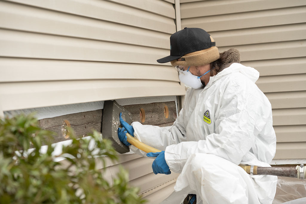a WxForce team member dressed in protective gear holding a hose that is being used to add insulation inside an exterior wall of a home