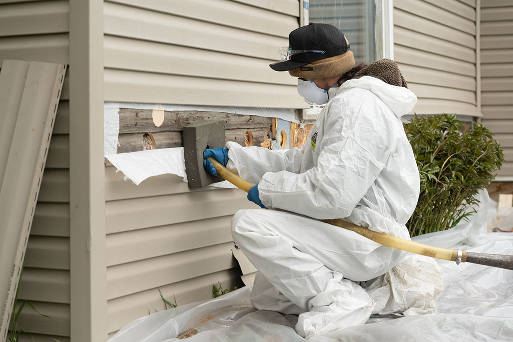a WxForce team member dressed in protective gear holding a hose that is being used to add insulation inside an exterior wall of a home