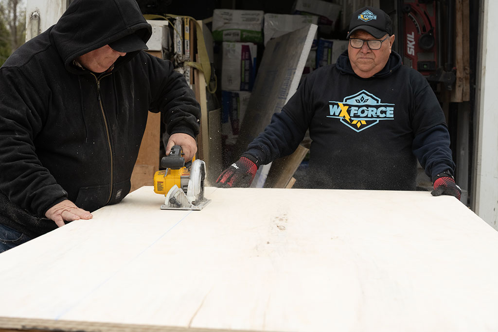 one WxForce team member is holding a piece of plywood while another team member is cutting it to size using a power hand saw