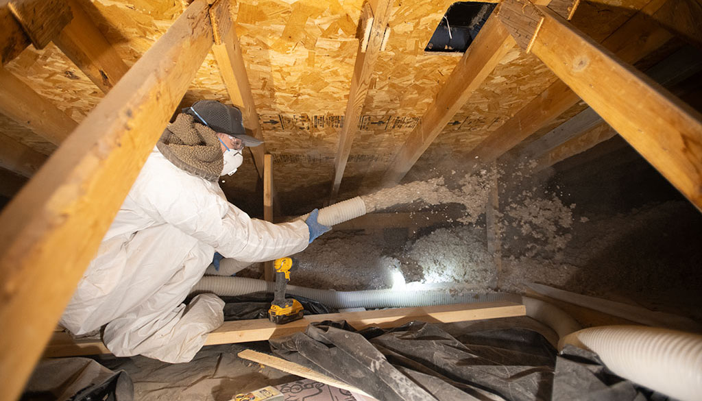 worker in an attic space holding a hose that is piping insulation into the space